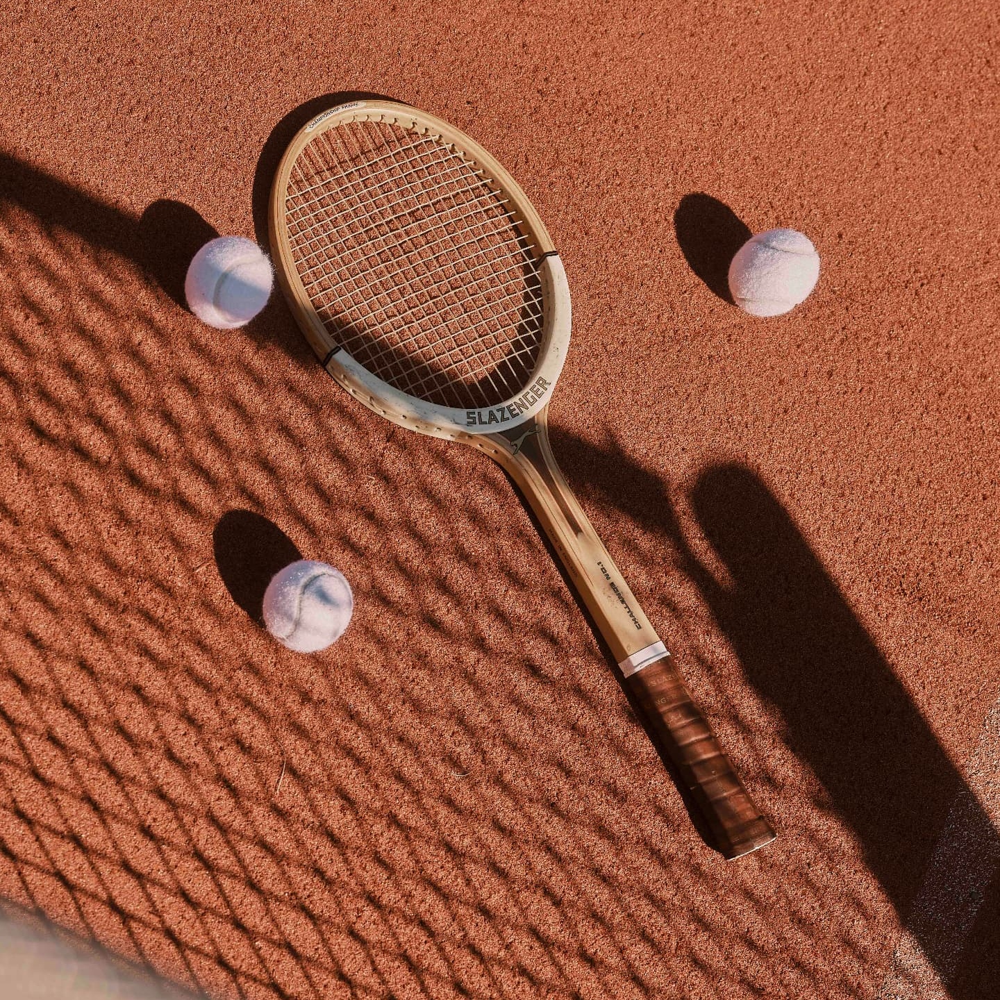 A tennis racket resting on red clay beside the net shadow, photographed by Jonathan Ducrest